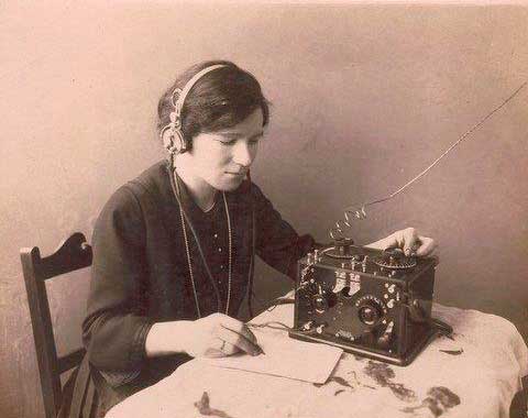Black and white photograph of a woman sitting at a table wearing headphones and operating a small machine. - click to view larger image