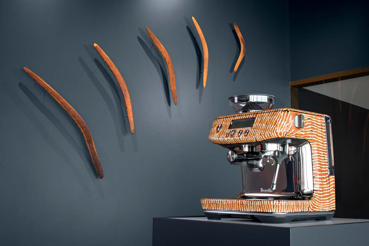 An espresso machine with a painted design on display in a gallery space. On the wall behind are wooden boomerangs on display. - click to view larger image