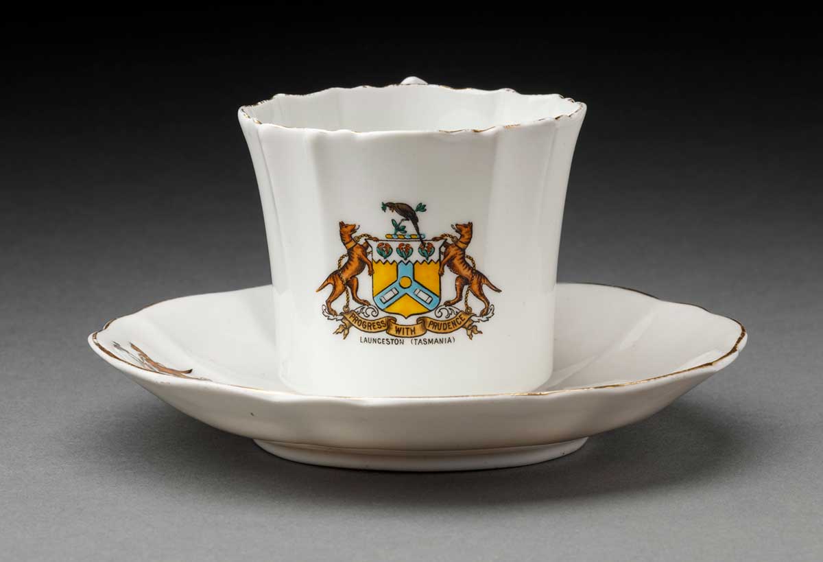 A white teacup and saucer set decorated with a coat of arms featuring two Tasmanian Tigers flanking a shield with the words 'PROGRESS WITH PRUDENCE / LAUNCESTON (TASMANIA)' below.