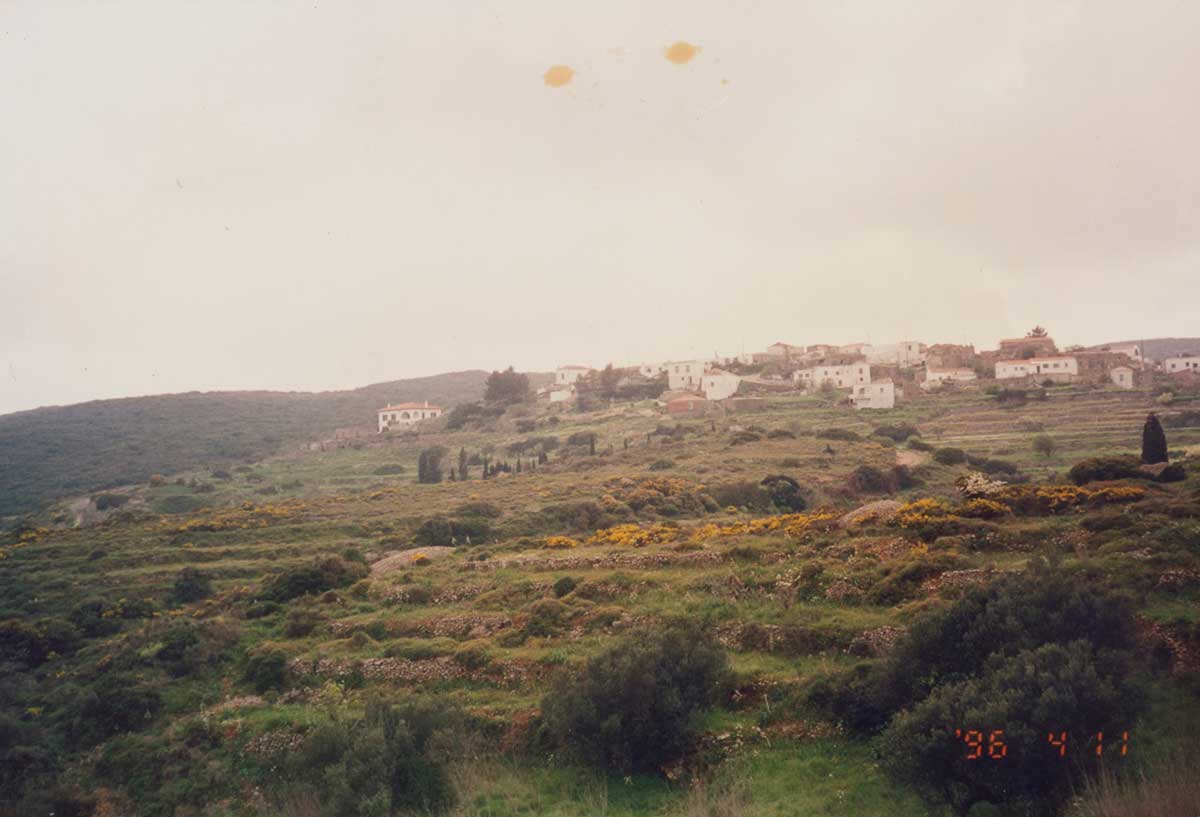 Landscape photograph of a small Greek village on a hill in a lush green landscape. The sky is overexposed and appears white, and the photo has a very light sepia appearance. There are two small stains at the top middle of the photograph. The numbers ‘96 4 11 are overlaid in the bottom right corner of the photograph, indicating the date the photograph was taken.