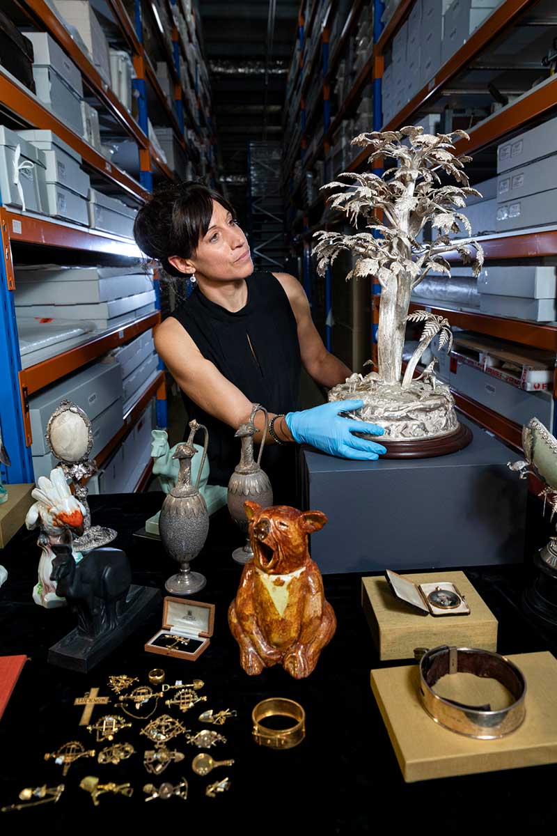 A woman wearing blue latex gloves is inspecting a collection of memorabilia. She is standing in an aisle of a large storage warehouse.