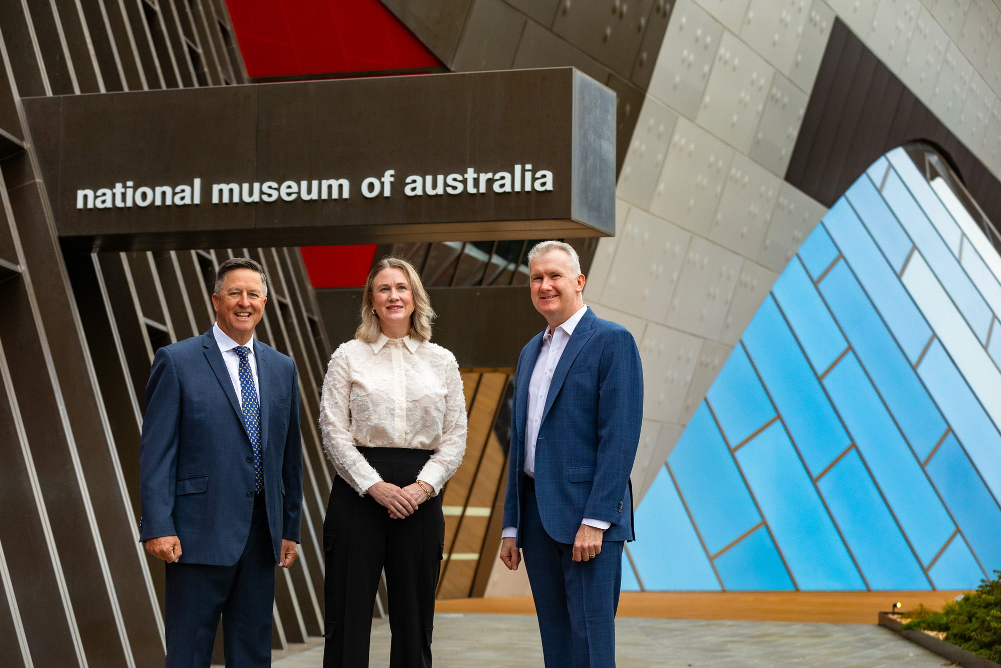 Three people stand outside the front entrance to the National Museum of Australia.