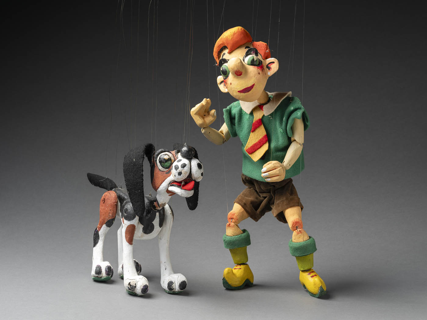 A photo of two string puppets. Right is a puppet of a boy named Nicky, he has bright orange hair and is wearing a green short sleeve shirt, brown shorts and a red and yellow tie. On his legs are green socks and yellow clog-like shoes. Left is a wooden puppet of a dog called Noodle. Noodle has a black, brown and white painted body, felt ears and green felt soles attached to its feet. Noodle has green painted eyes and a red felt tongue. 