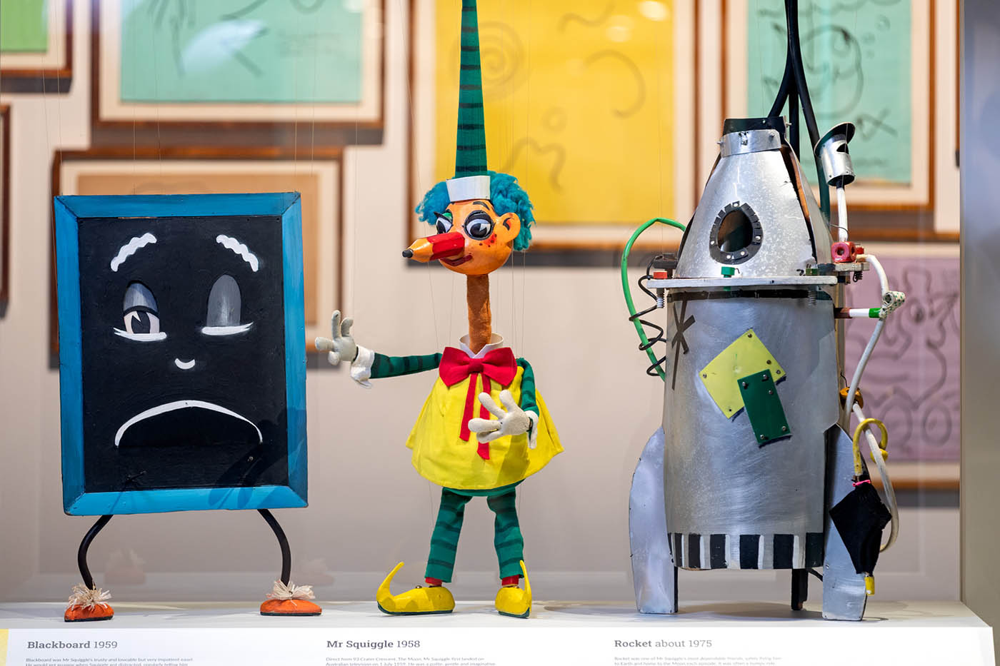  Museum case displaying three puppets, from left, a blackboard with feet and a face, a man with a pencil for a nose and a bright yellow frock and a rocket with three fins and a small viewing hole in the nose cone. 