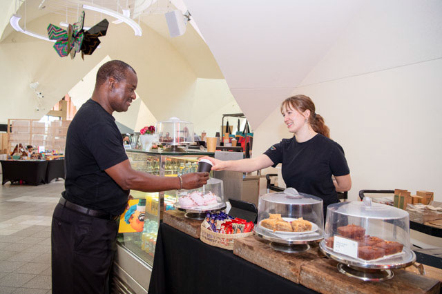 A woman hands a man a takeaway coffee cup at a stall with baked goods and chocolates on display. - click to view larger image