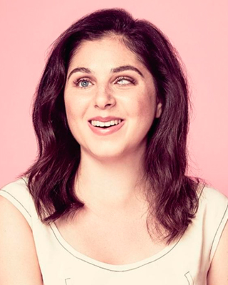 A photograph of Nas Campanella. NAs is smiling, she has brown slightly wavy hair and is in front of a pink backdrop.