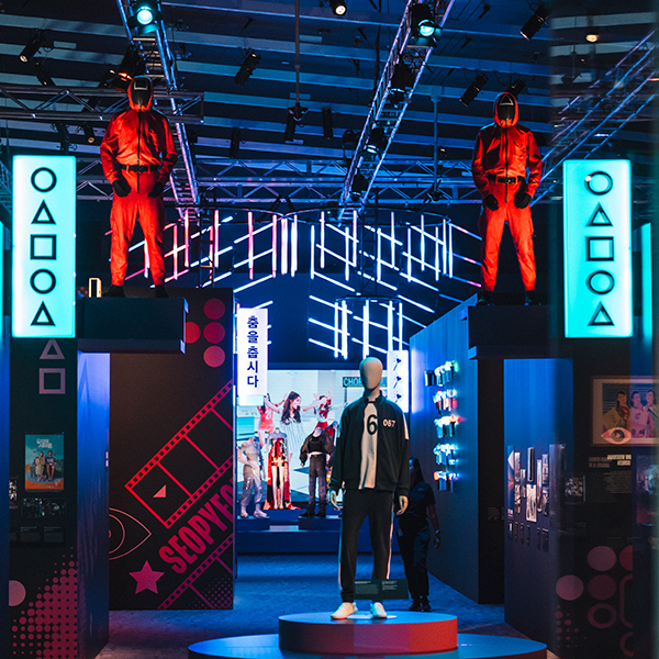 A photograph of the Hallyu exhibition, focusing on the Squid game tracksuits on mannequins. The mannequins are amongst the colourful exhibition space which has lots of lights and screens.