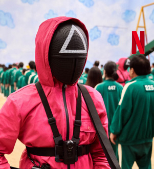 A man in a hooded pink tracksuit, with a black face mask with a white triangle symbol on the forehead, stands amid a field of people, some wearing green tracksuits, unmasked, and others wearing black face masks and pink tracksuits.