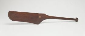Dance paddle made of wood with an almost rectangular and rounded blade (that is missing on one side) and a semi-circular knob at the end of the handle.