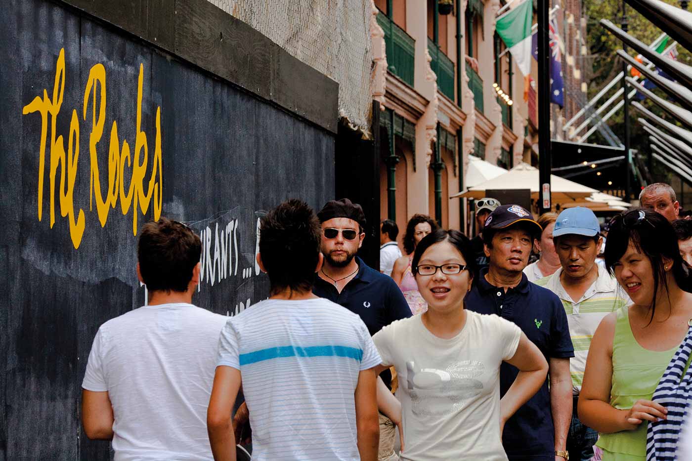 Urban street view showing people walking alongside a painted black wall. 'The Rocks' has been handpainted in a yellow script at the top left. 