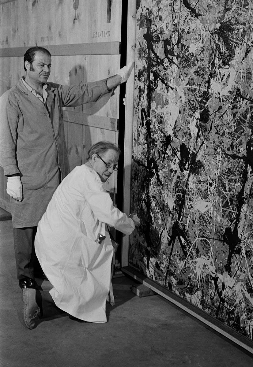 Black and white photo of two men in protective coats, one standing, one crouching, by Blue Poles, part of which is visible. Three of its distinctive poles can be seen. These are thick, dark more or less vertical lines of splattered paint. These have been painted over a splattered arrangement of different coloured paint. - click to view larger image