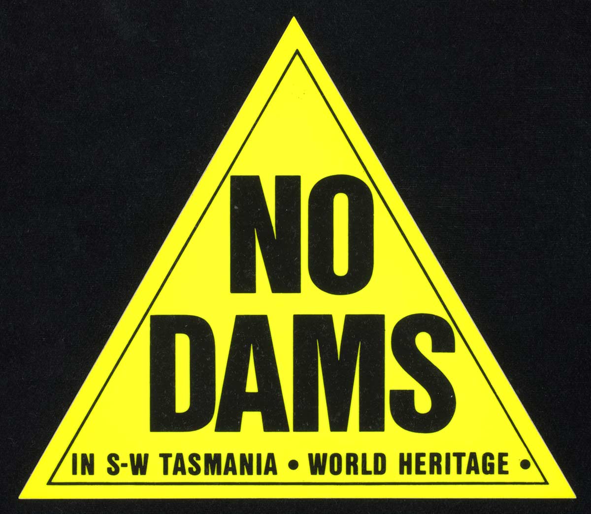 Yellow triangle on black background. Within the triangle are the words NO DAMS in capital letters. Beneath these are the words ‘IN S W TASMANIA. WORLD HERITAGE. - click to view larger image