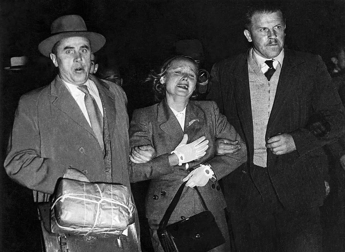 Photo taken at night showing distressed woman flanked by two men in suits holding her arms.