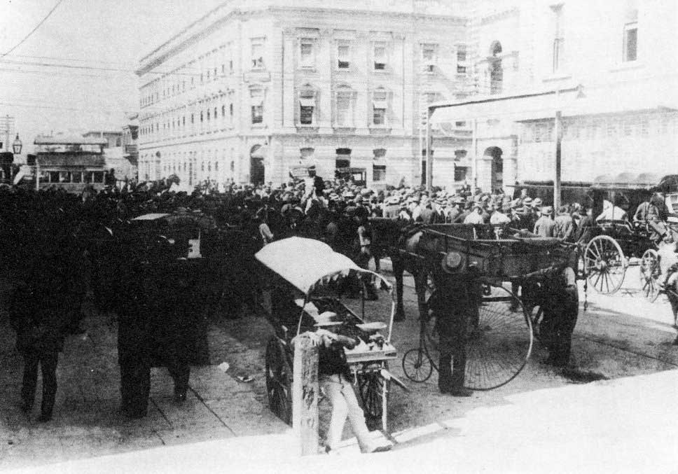 Black and white photo of a large crowd of people on a street with horse and carriage.