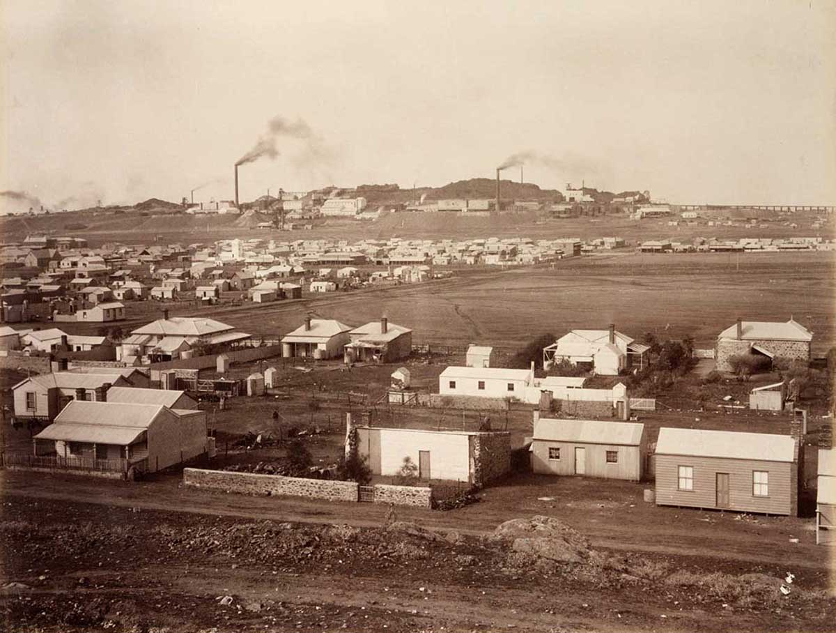 Black and white photograph of a township. In the distance a factory with smoke stacks can be seen.