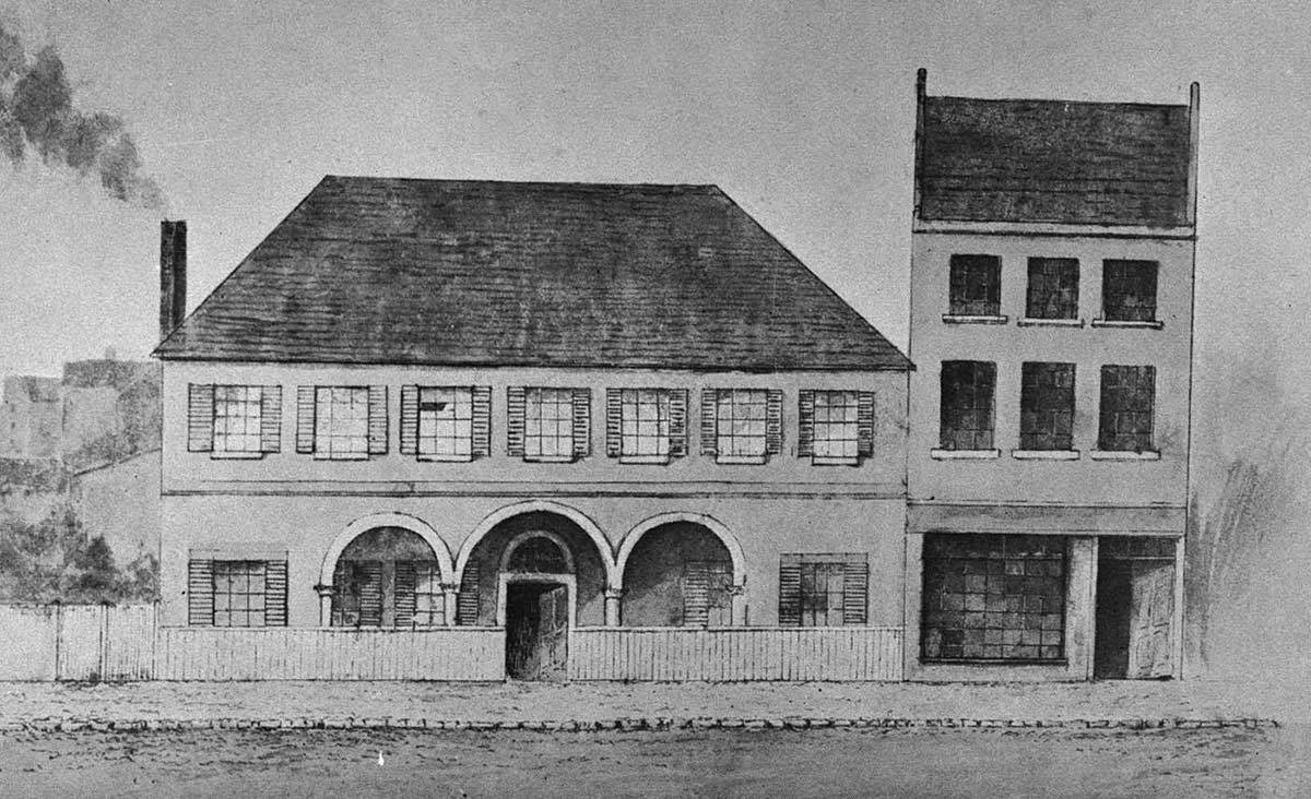 Black and white drawing or lithograph of a two-storey building with arched façade and a smoking chimney. Adjoining it is a three-storey terrace with a shop at the bottom. There are no people visible.