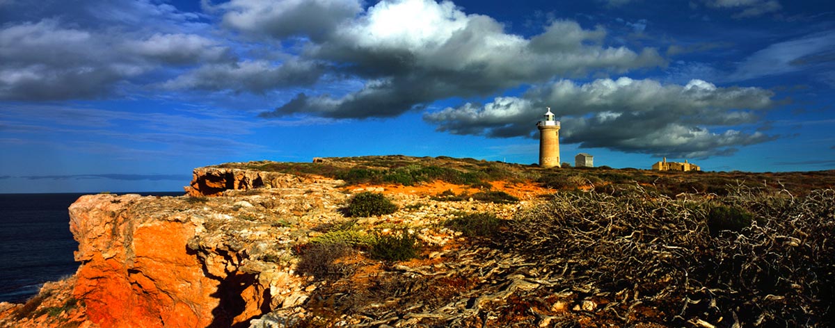 Colour photo of a piece of coastline showing cliffs of orange rock and a small lighthouse in the distance.