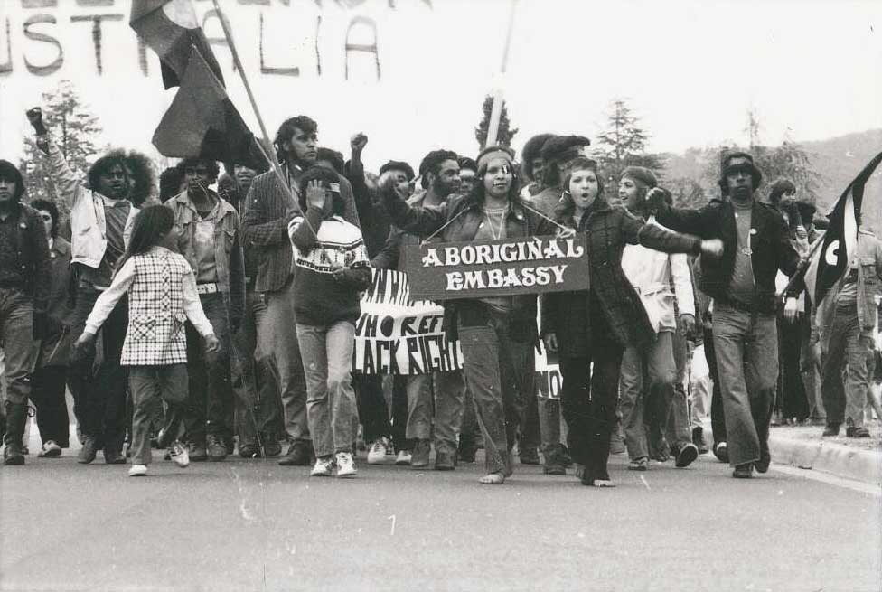 Aboriginal people march, holding flags and banners. A woman in the front has a placard around her neck reading ‘Aboriginal Embassy’.