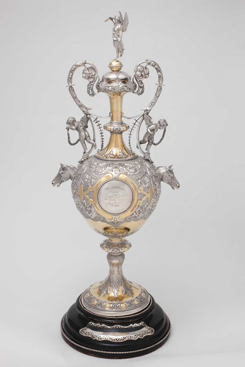 A silver-gilt chased cup with gold leaf, consisting of a base, cup and lid. The cup is decorated with silver foliage designs and two silver horse heads on either side. A central shield is inscribed with the words ''MELBOURNE / CUP / 1866 / WON BY / MR JOHN TAIT'S / BLK. H. The Barb / 3 years'. Two cherubs are attached to the ornate handles at the top of neck, and an angel holding a wreath sits on the top of the lid. The circular base is painted black. - click to view larger image