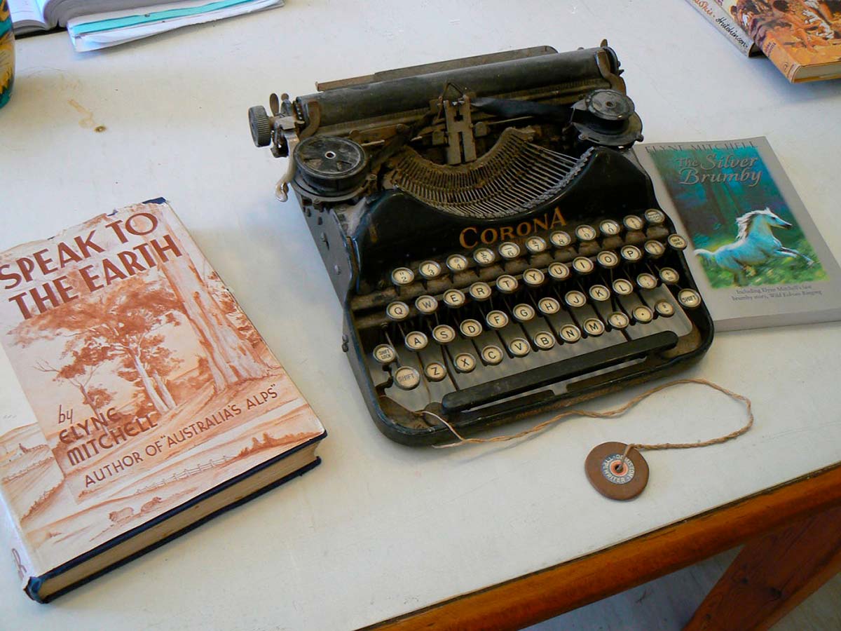 An old-fashioned typewriter on a desk. The typewriter has round keys and the word 'Corona' on the front of the mechanism casing. A piece of string is tied to the front of the typewriter; a disc with a hole in it is attached to the other end of the string. Two books are next to the typewriter. The book at left is called Speak To The Earth while the book at right is called The Silver Brumby.