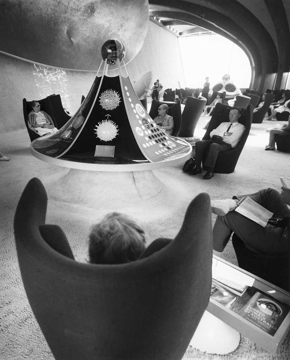 Black and white image showing people sitting in high-backed winged chairs. A circular display case, which rises to a peak is also visible.