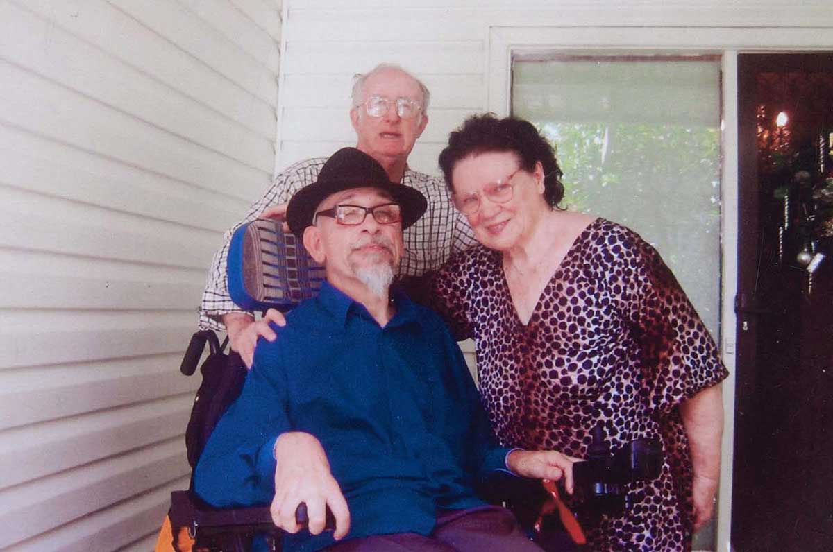 A man sits in a wheelchair. A man standing behind rests his right hand on the seated man's shoulder. A woman leans in to put her right arm around the seated man. - click to view larger image