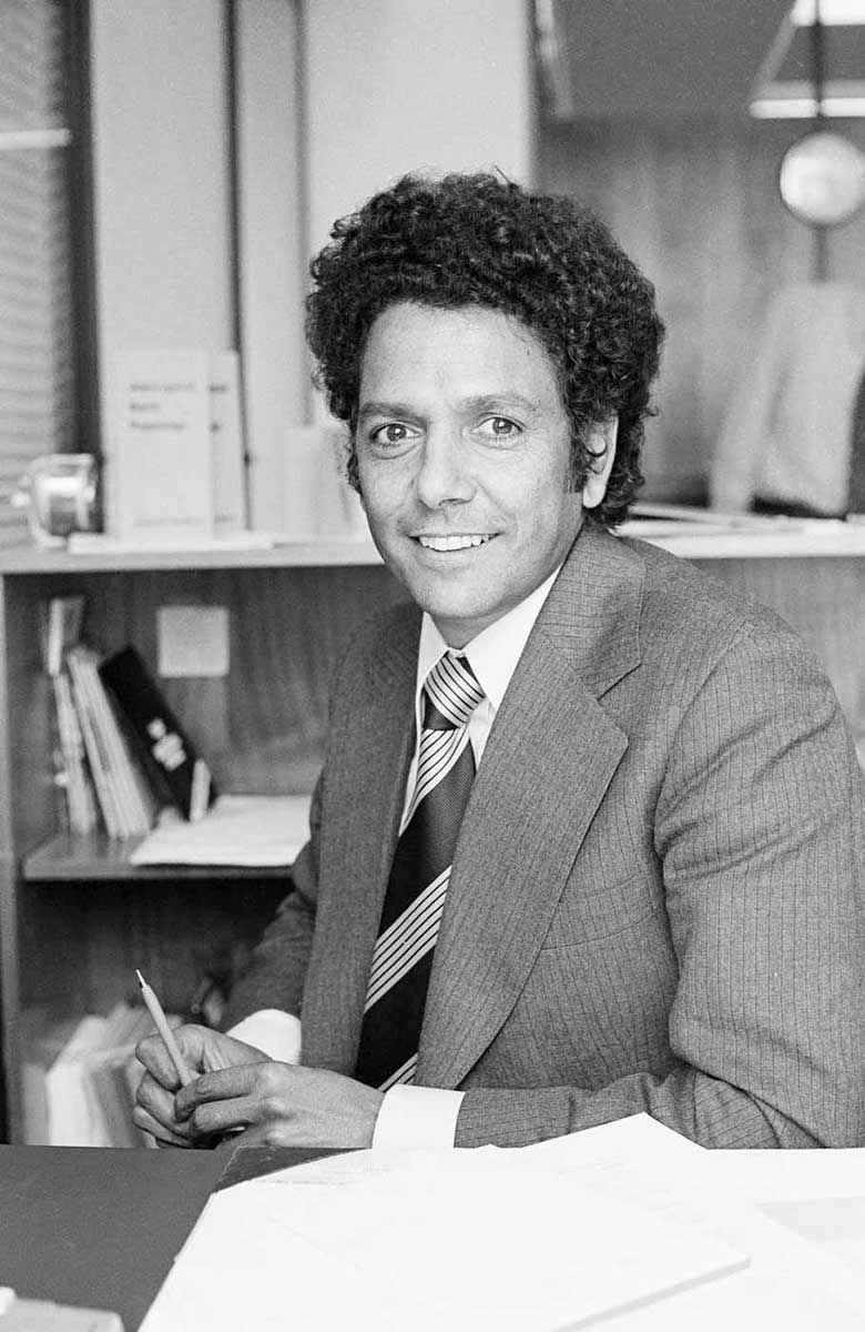 Photograph of John Moriarty sitting at a desk with books on the shelves behind and a pen in his right hand. - click to view larger image