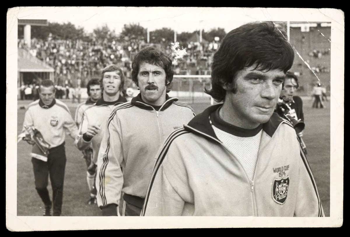 Black and white photograph showing five men walking on a grass field. The man at front right is wearing a tracksuit top with the Australian coat of arms on the right breast and the words 'World Cup 1974' embroidered above. Behind him are four other three other men wearing tracksuit tops. A partially obscured man carrying a camera is visible on the right and crowds of people in tiered seating are in the background.