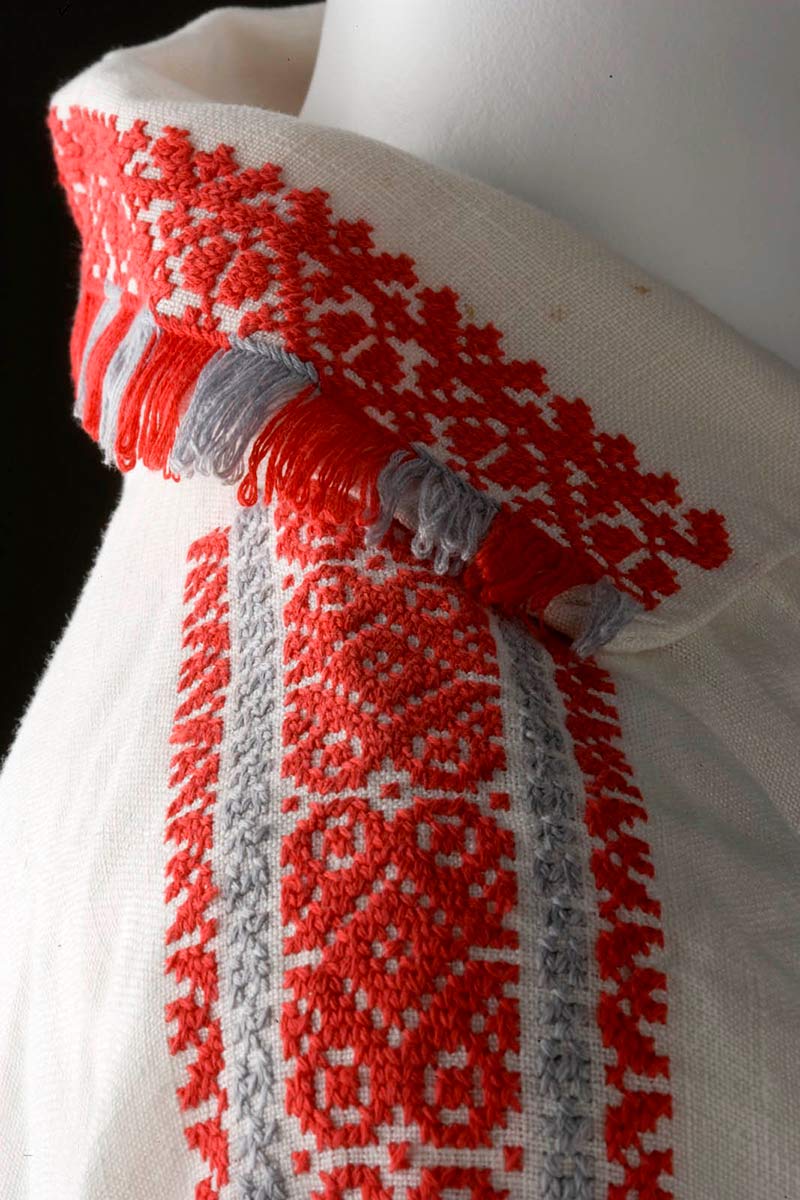 Side view showing a section of white linen shirt with the collar and sleeve embroidered in orange and grey thread. The collar is tessellated with the same thread. - click to view larger image