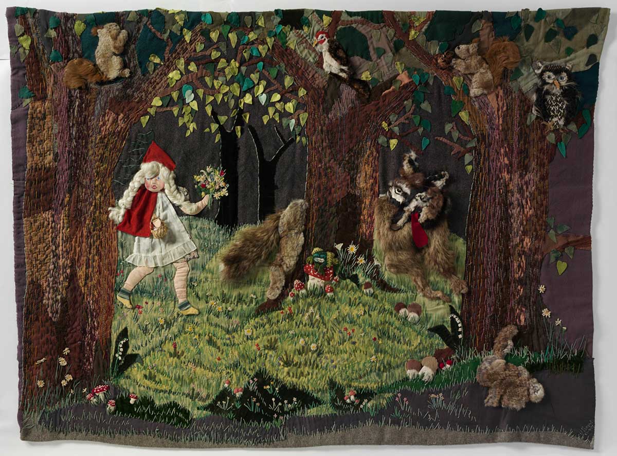 Wall hanging depicting Little Red Riding Hood walking through a forest. A wolf appears from behind a tree, with his red tongue hanging out the side of his mouth. A variety of fabrics including felt and fur have been sewn onto a grey blanket to form the scene, which also shows an owl, a bird, a squirrel and a rabbit. Some of the elements are padded using a technique called stumping, and they have a three-dimensional appearance.
