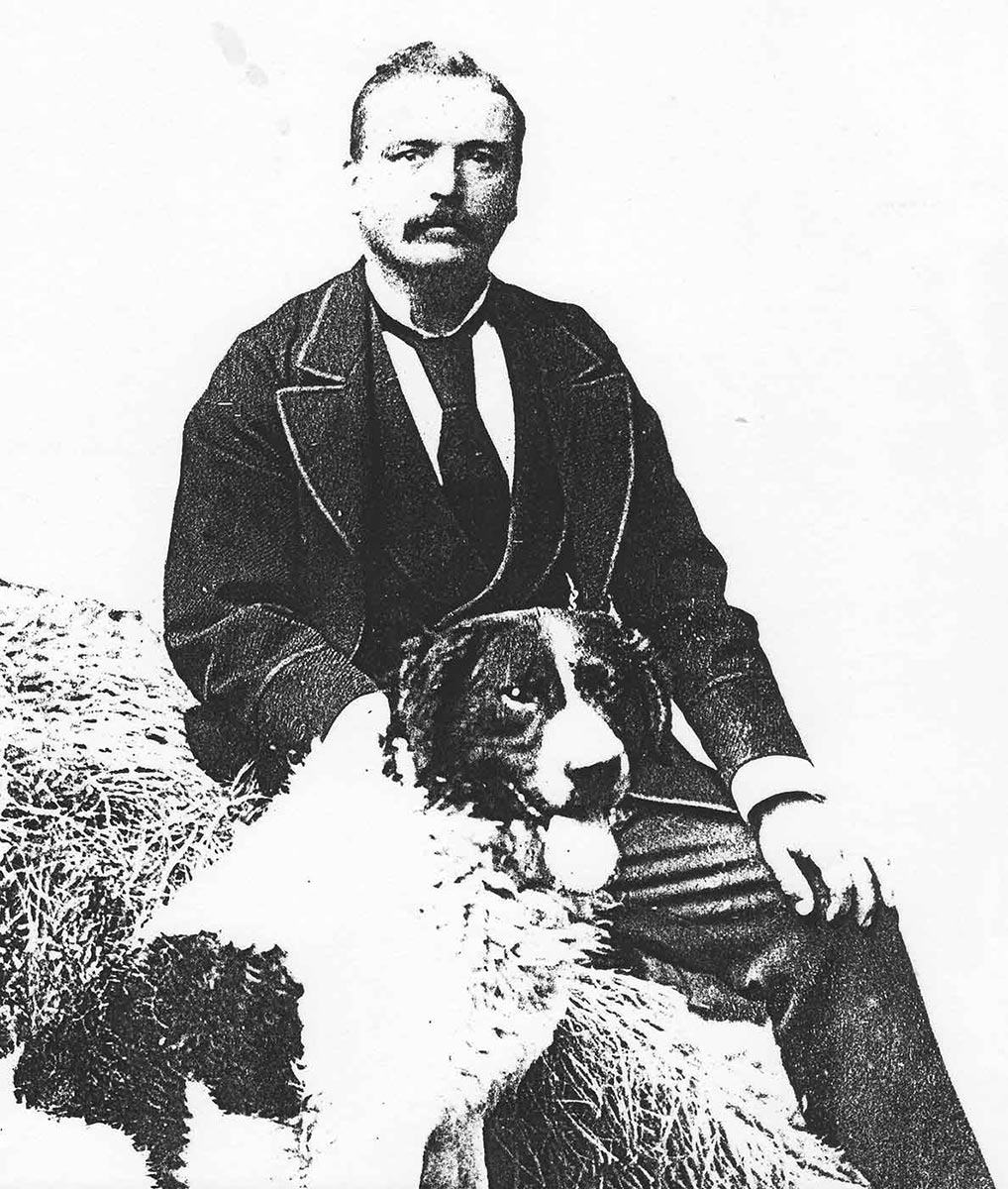 Black and white studio portrait showing a man seated on a hay bale, with a large two-toned dog sitting at his right knee. - click to view larger image