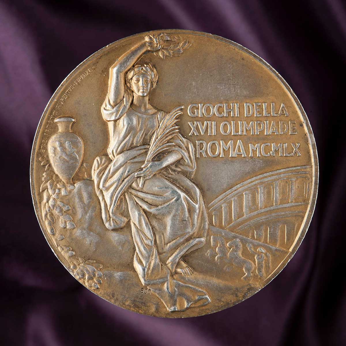 Yellow-coloured circular medal with a classical image showing a woman dressed in a robe, sitting cradling a small branch in her right arm and holding a laurel above her head with her left. An urn apears to her left and part of an arched bridge to her right. The medal has been inscribed with the text 'GIOCHI DELLA / XVII OLIMPIADE / ROMA MCMLX'.