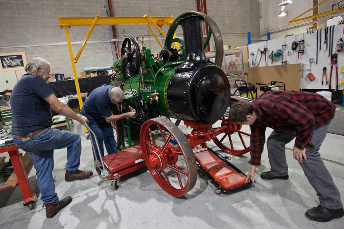 A colour photograph of three men removing trolleys from under a large steam engine. - click to view larger image
