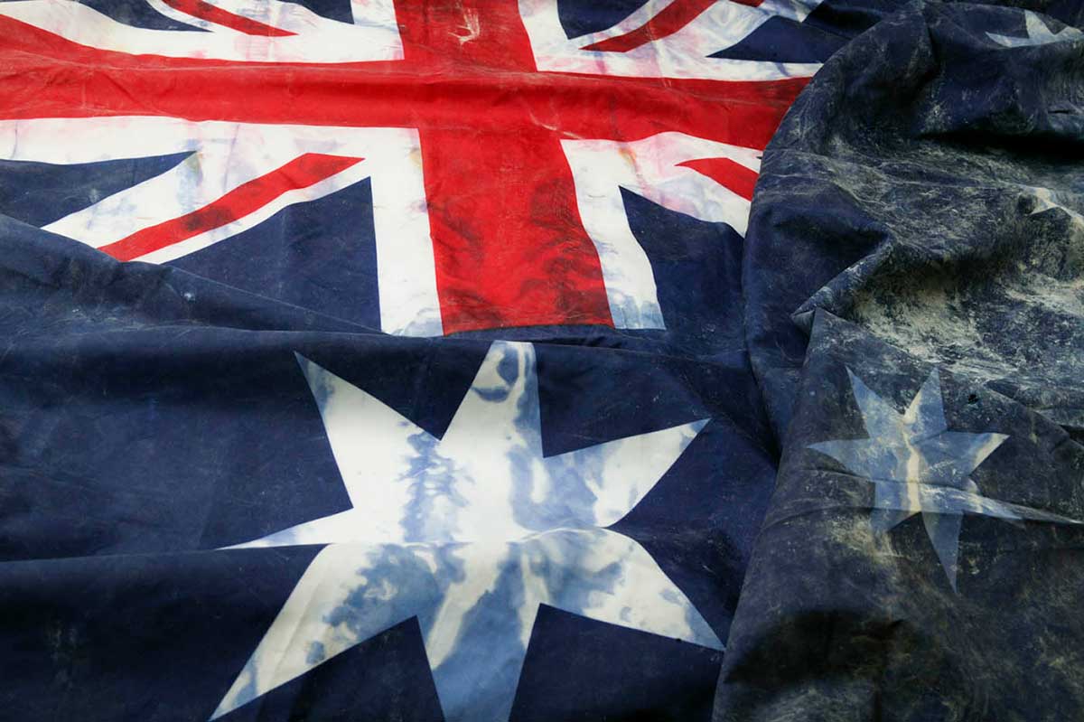 Top left section of an Australian flag with stains, tears and burns. The Union Jack is visible, along with the large seven-pointed star. The section bearing the Southern Cross is quite crumpled.