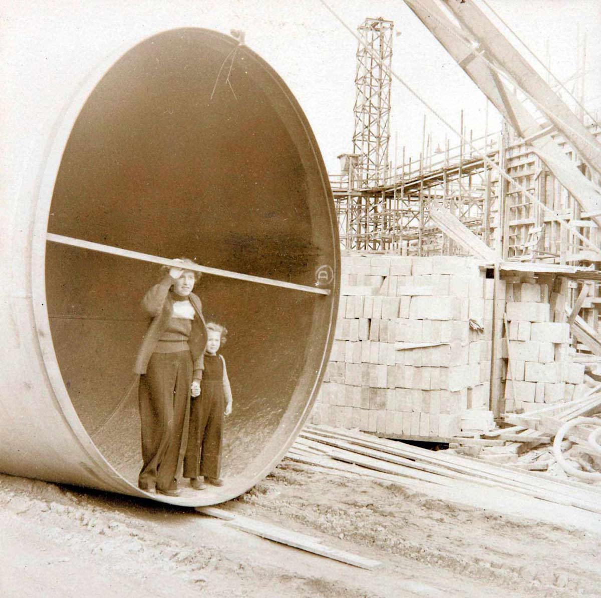 Photograph of a woman and a young girl standing inside the open end of a very large pipe at a building site. - click to view larger image