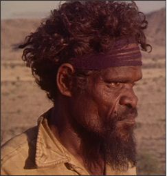 Side profile of a man with dark curly hair and a beard, and wearing a maroon headband and yellow open-collared shirt.