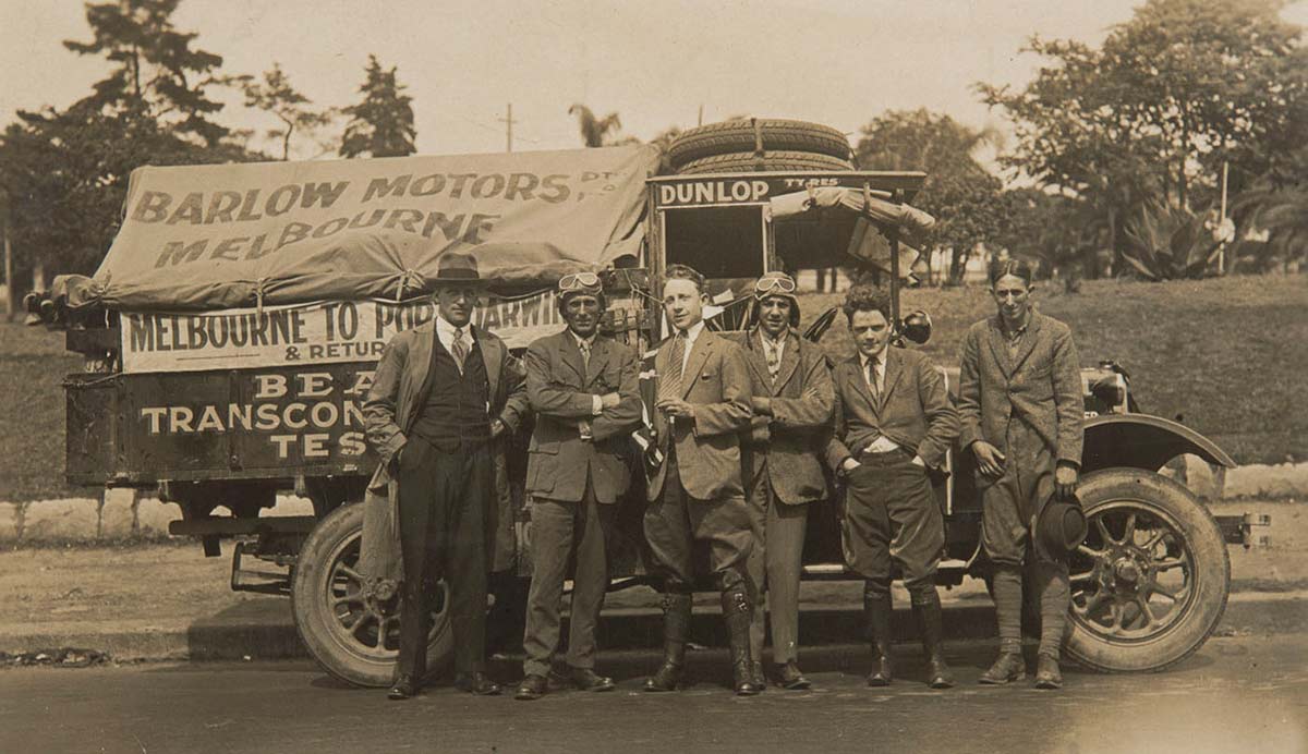 Black and white image showing six men standing in front of a truck which has a canvas cover at the back bearing the words 'BARLOW MOTORS / Melbourne'. All the men wear suits. Those standing second and fourth from left also wear leather flying-style caps and goggles on their heads.