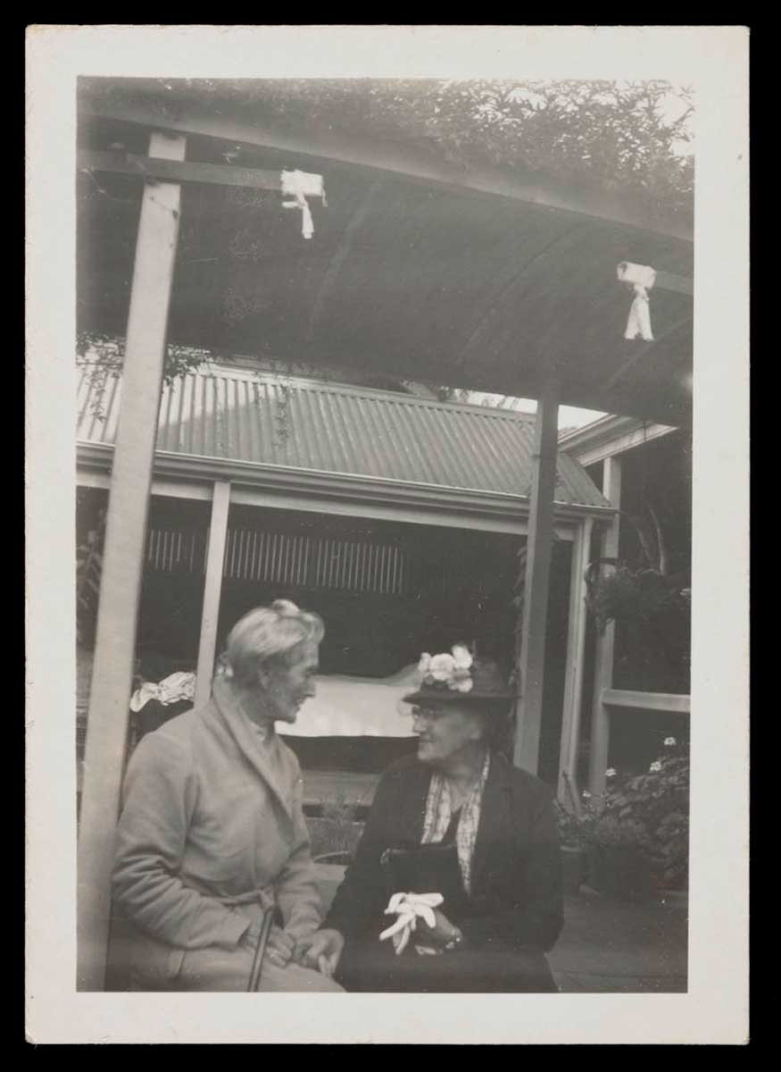 Black and white photograph showing two women sitting under a garden structure with a corrugated iron roof. The woman on the right holds the hand of the woman on the left. - click to view larger image