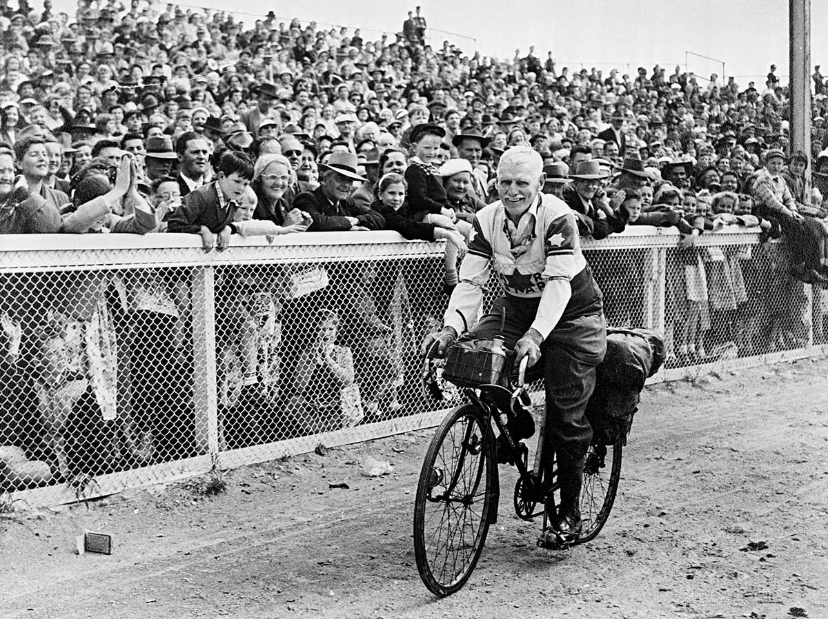 A black and white photo of an older man sitting on a bike with a crowd behind him.