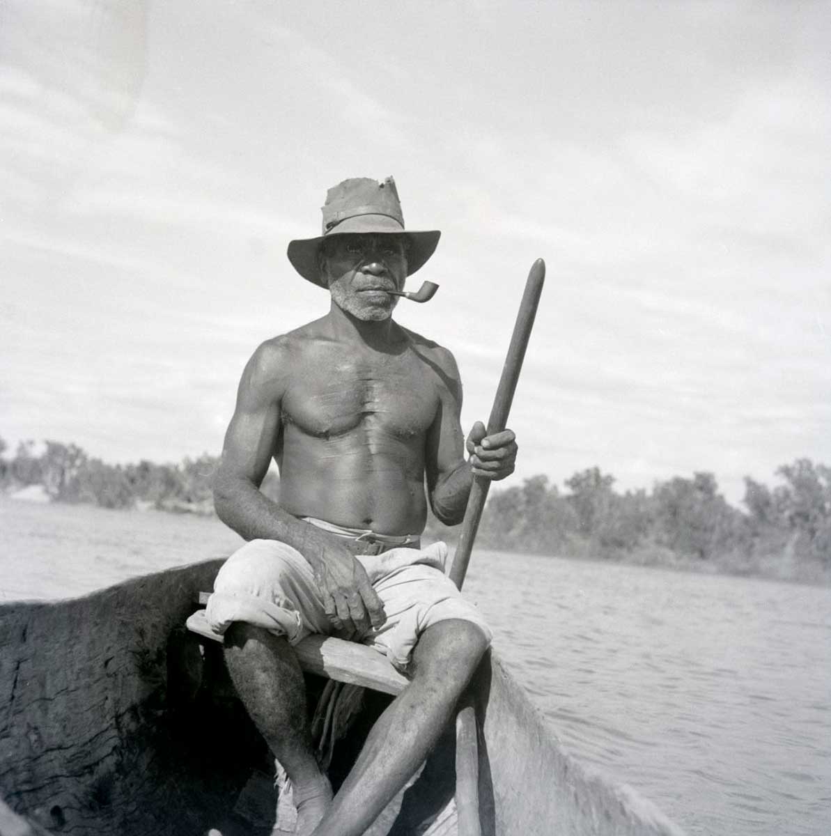 A black and white photographic negative that depicts an Aboriginal man wearing a hat with a pipe in his mouth sitting in a canoe on a river. - click to view larger image
