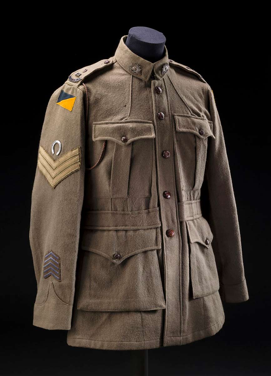 A tunic from a light horse uniform. It is khaki green in colour and has four pockets at the front. It has five brown buttons down the centre and one on each pocket flat. The collar has a rising sun 'AUSTRALIAN / COMMONWEALTH / MILITARY SERVICES' badge on each side. The top of each shoulder has metal '9 / LH / AUSTRALIA' on them. The top of each arm shows a patch that has a green rectangle at the top and a gold one at the bottom to form a rectangle. The proper right arm then has a small metal horse shoe above three khaki strips in a downward pointing arrow. The lower part of the proper right sleeve has four blue stripes in an upward pointing arrow. - click to view larger image