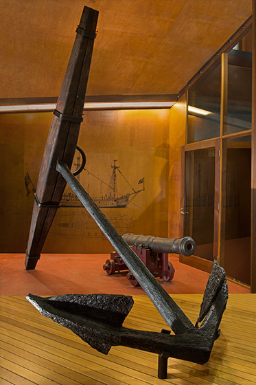 Museum display showing a metal ship's anchor with wooden stock. A cannon and diagram of a ship are also visible.
