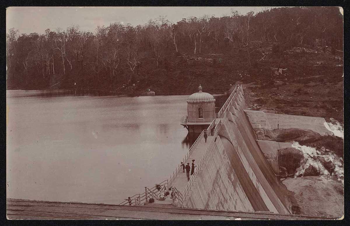 A sepia photo taken from above a concrete dam wall. Several people stand on the wall, looking at the water on one side, and the spillway on the other. - click to view larger image