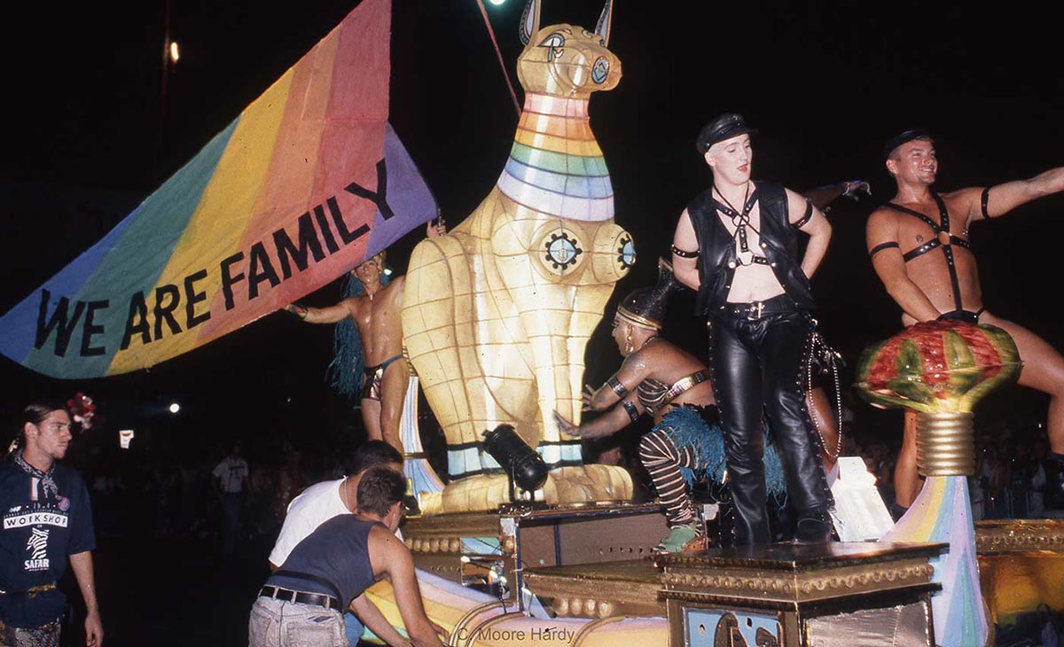 Men in leather pose next to a gigantic cat figure with a rainbow banner stating ‘We are family’. - click to view larger image