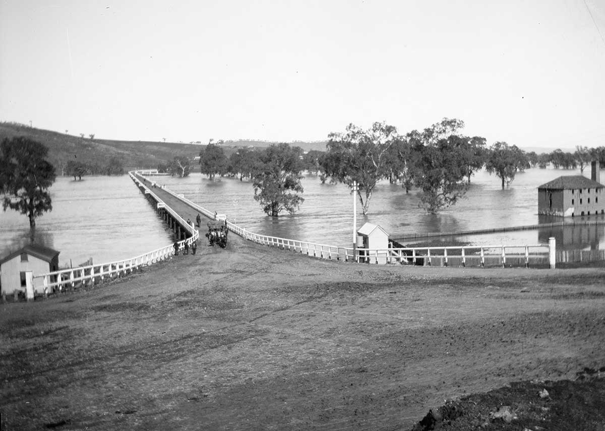 Photo shows swollen river with bridge almost covered in water as well as building and trees surrounded by water.