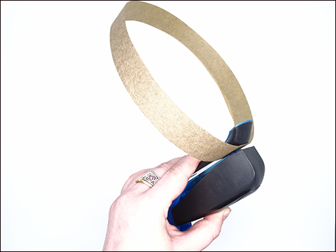 The top and back part of someone's hand holding a black stapler to a bronze coloured strip of cardboard in the shape of a headband.