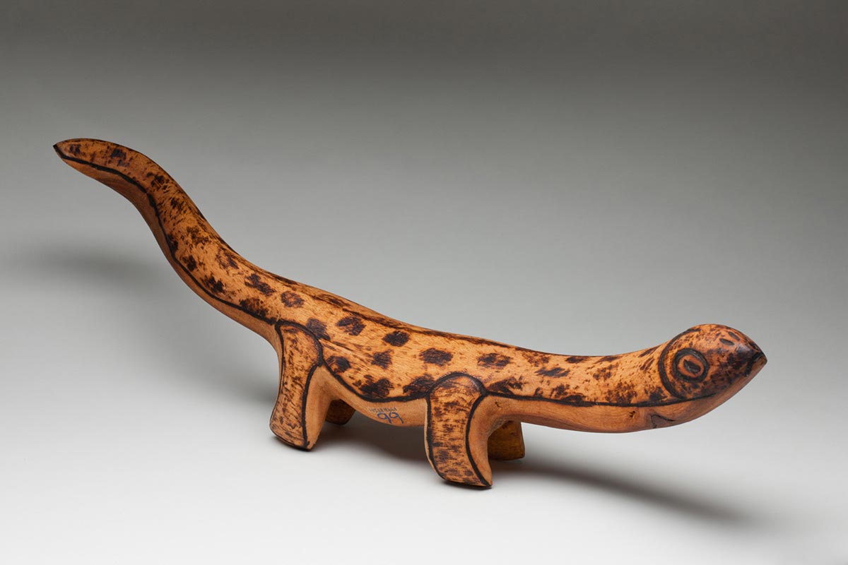 Wooden lizard sculpture standing upright on four legs with a thick neck and head pointed upwards and tilted slightly to the right, and a long tail which points upwards and twists to the right with burnt pokerwork designs overall. - click to view larger image