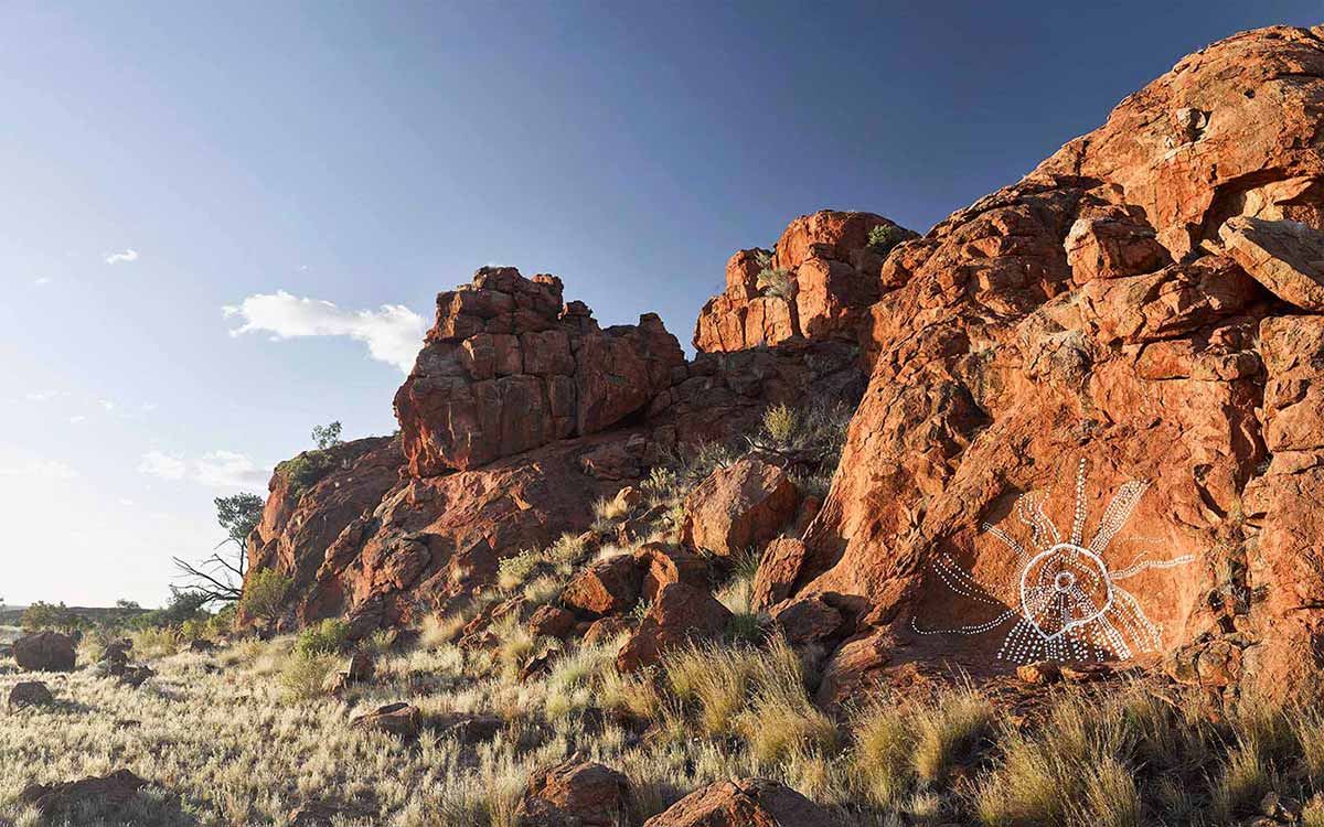 Landscape photography showing red rocks beside a grassy plain. Part of the largest rock in the right foreground has been painted with white dots extending from a central white circle. - click to view larger image