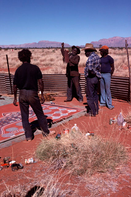 Uta Uta Tjangala raises his arm, watched by three men.