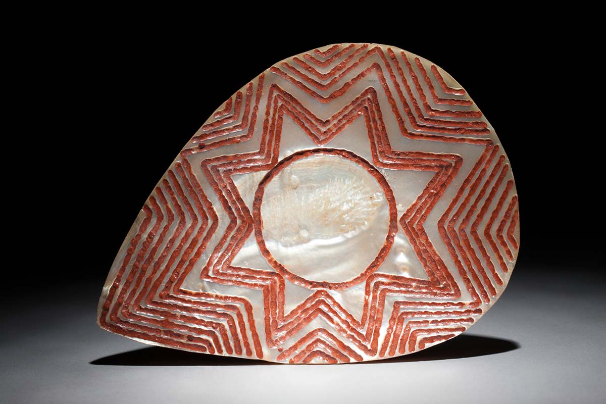A pearl shell which has been incised with a geometric pattern around a central circle and star shape. The incised patterns have been painted red. - click to view larger image